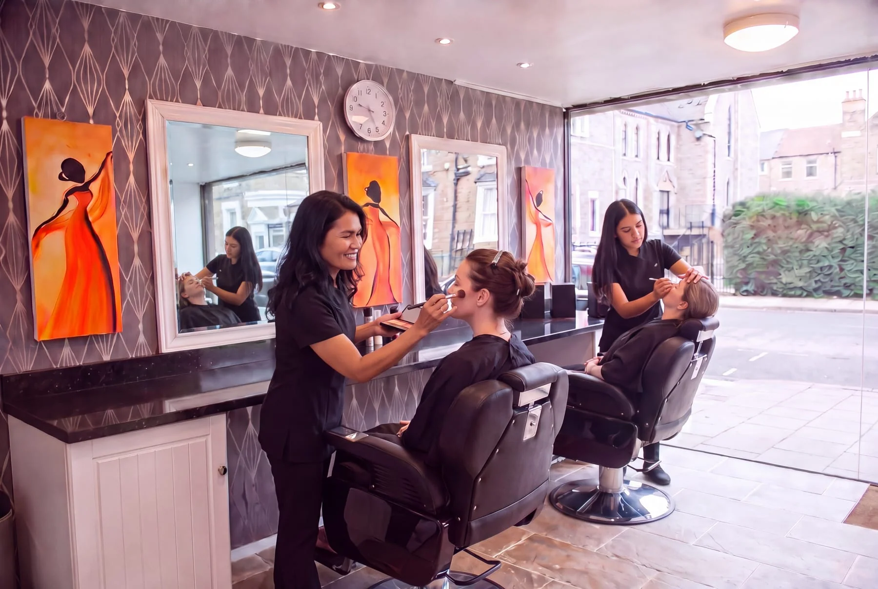 A salon styling room with two clients in black styling chairs, one having her make-up applied and the other having her eyebrows shaped, in front of a black counter, white-framed mirrors and orange canvases on patterned grey wallpaper, beside a full-height shopfront window